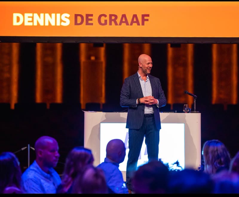 Dennis de Graaf in krijtstreeppak op een podium, met publiek als donkere rand