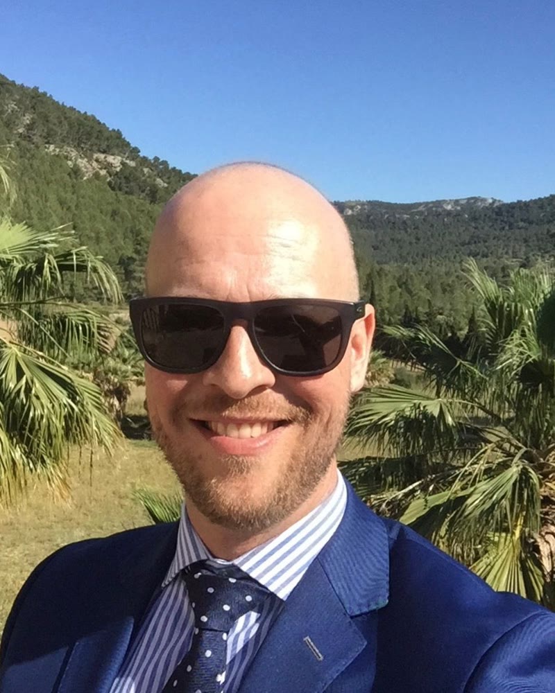 Dennis in een blauw pak met zonnebril, palmen en bergen achter hem