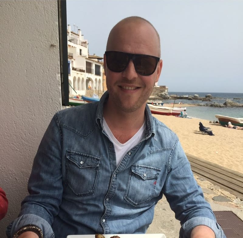 Dennis aan een tafel aan zee, met zonnebril, bootjes en strand achter hem